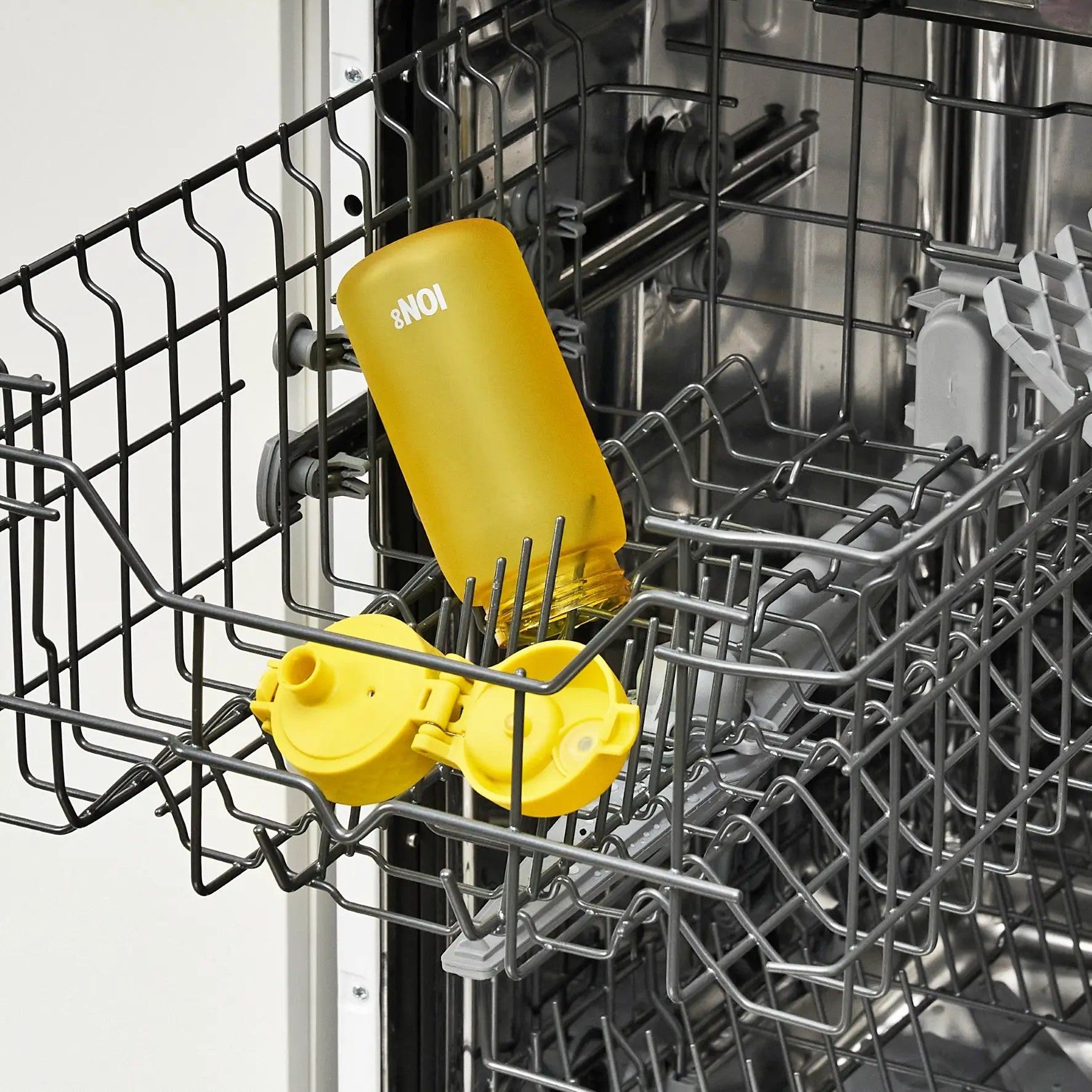 A yellow 8NOI water bottle and its lid are being washed inside a dishwasher's top rack.