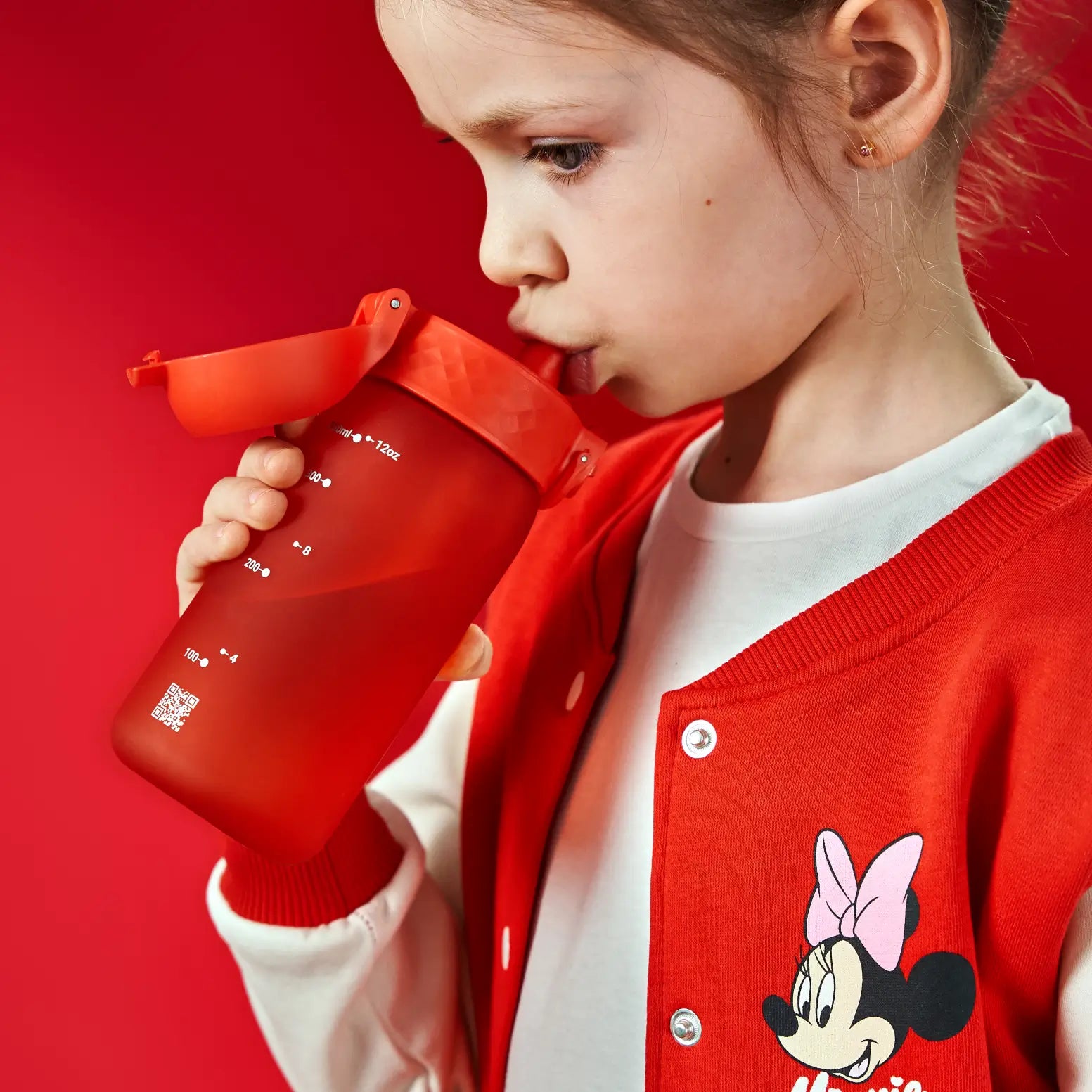 A child drinks from a red, graduated water bottle with a flip top. The bottle shows measurements in milliliters and ounces. The child wears a red jacket with Minnie Mouse on it. ml-12oz. 100-4. 200-8.