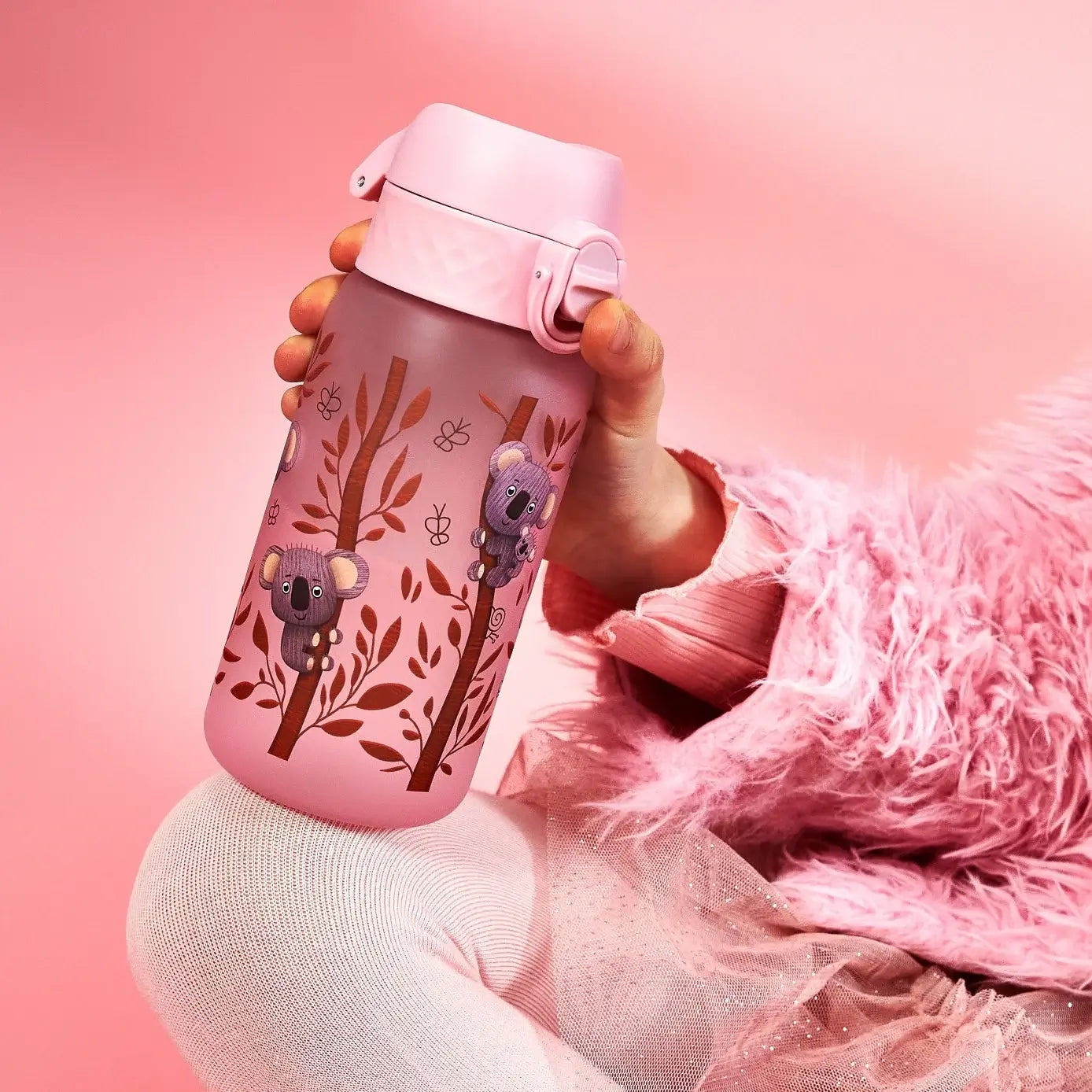 A pink children's water bottle, featuring koala illustrations, is being held by a child wearing a pink fuzzy jacket, against a pink background.