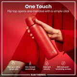 A red, slim thermal water bottle is being held; its flip top opens one-handed with a simple click; against a red background.
