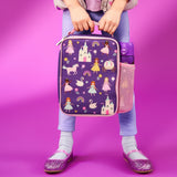 Child holding purple lunchbox featuring princesses and castles with a water bottle in a side pocket standing against a pink background wearing pastel clothes and sparkly shoes