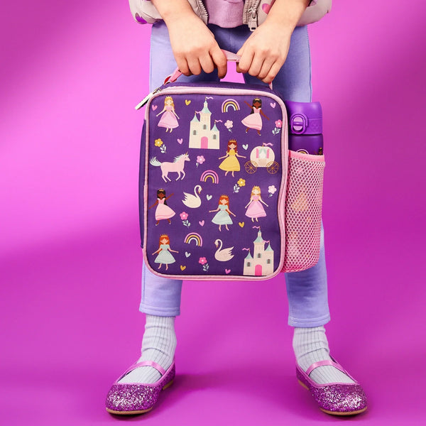 Child holding purple lunchbox featuring princesses and castles with a water bottle in a side pocket standing against a pink background wearing pastel clothes and sparkly shoes
