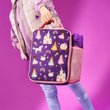 A child carries a purple lunchbox with princesses, castles, and unicorns, alongside a purple water bottle, wearing purple clothes against a solid purple background.