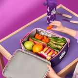A lunchbox with fruit, sandwich, snacks, and carrots being picked up, rests on a purple table with a purple water bottle featuring unicorn and castle designs nearby