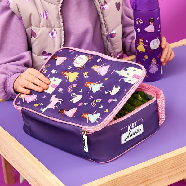 A child's purple lunchbox with princess and unicorn patterns is being opened. It's on a wooden table with a matching water bottle. The label reads ION8 Amelia.