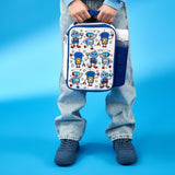 Lunchbox featuring colorful robot designs is held by a person wearing blue sneakers and jeans standing against a bright blue backdrop with a water bottle in a side pocket