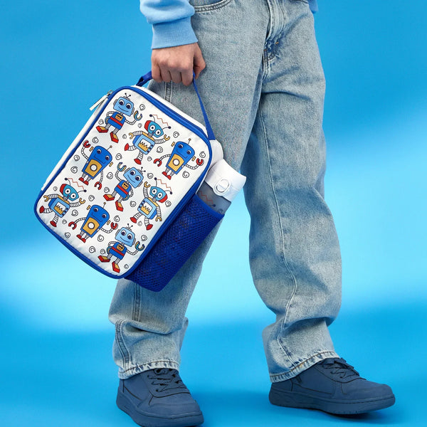A person carries a robot-themed lunchbox with a handle beside a water bottle in a blue mesh pocket wearing light blue jeans and sneakers against a blue background.