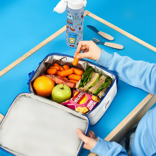 A packed lunchbox contains sandwiches, an apple, an orange, Walkers crisps, and carrot sticks. One hand picks up a carrot in a blue setting with a water bottle nearby.