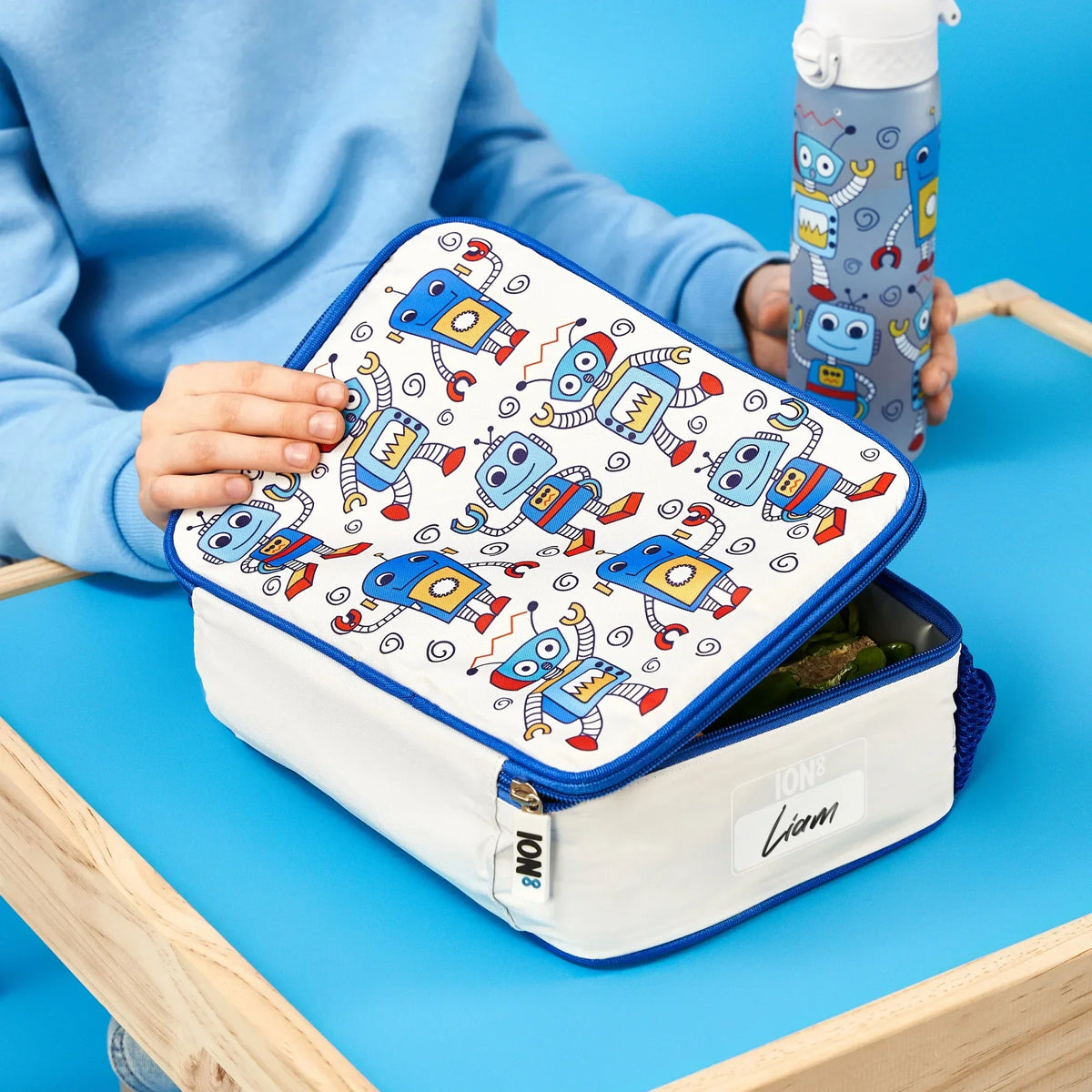 A lunchbox with blue robot designs is being opened by a child in a blue sweatshirt holding a matching bottle at a wooden table on a blue background. Text: ION8 Liam