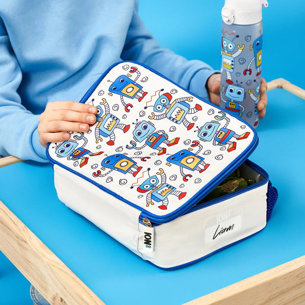 A lunchbox with blue robot designs is being opened by a child in a blue sweatshirt holding a matching bottle at a wooden table on a blue background. Text: ION8 Liam