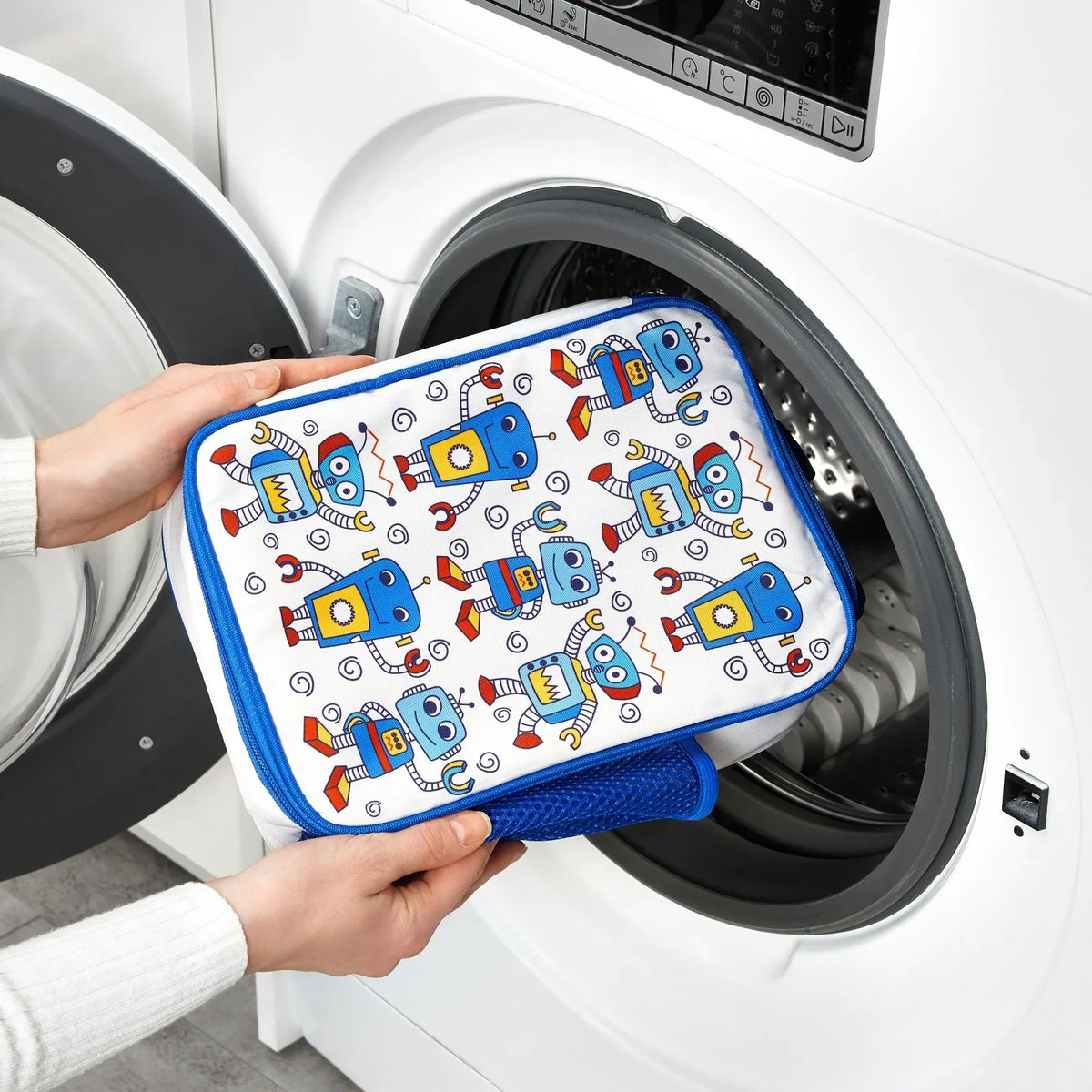 Hands are placing a colorful pouch decorated with blue robots into an open front-loading washing machine in a bright laundry room.