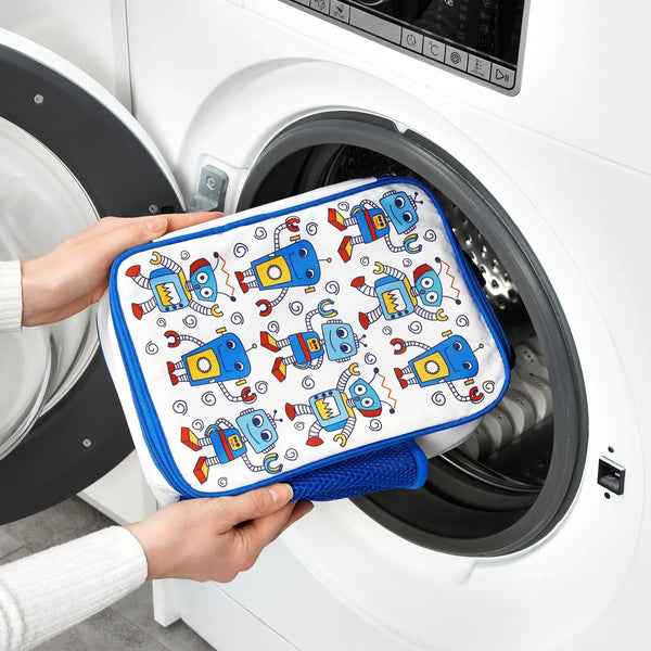 Hands are placing a colorful pouch decorated with blue robots into an open front-loading washing machine in a bright laundry room.