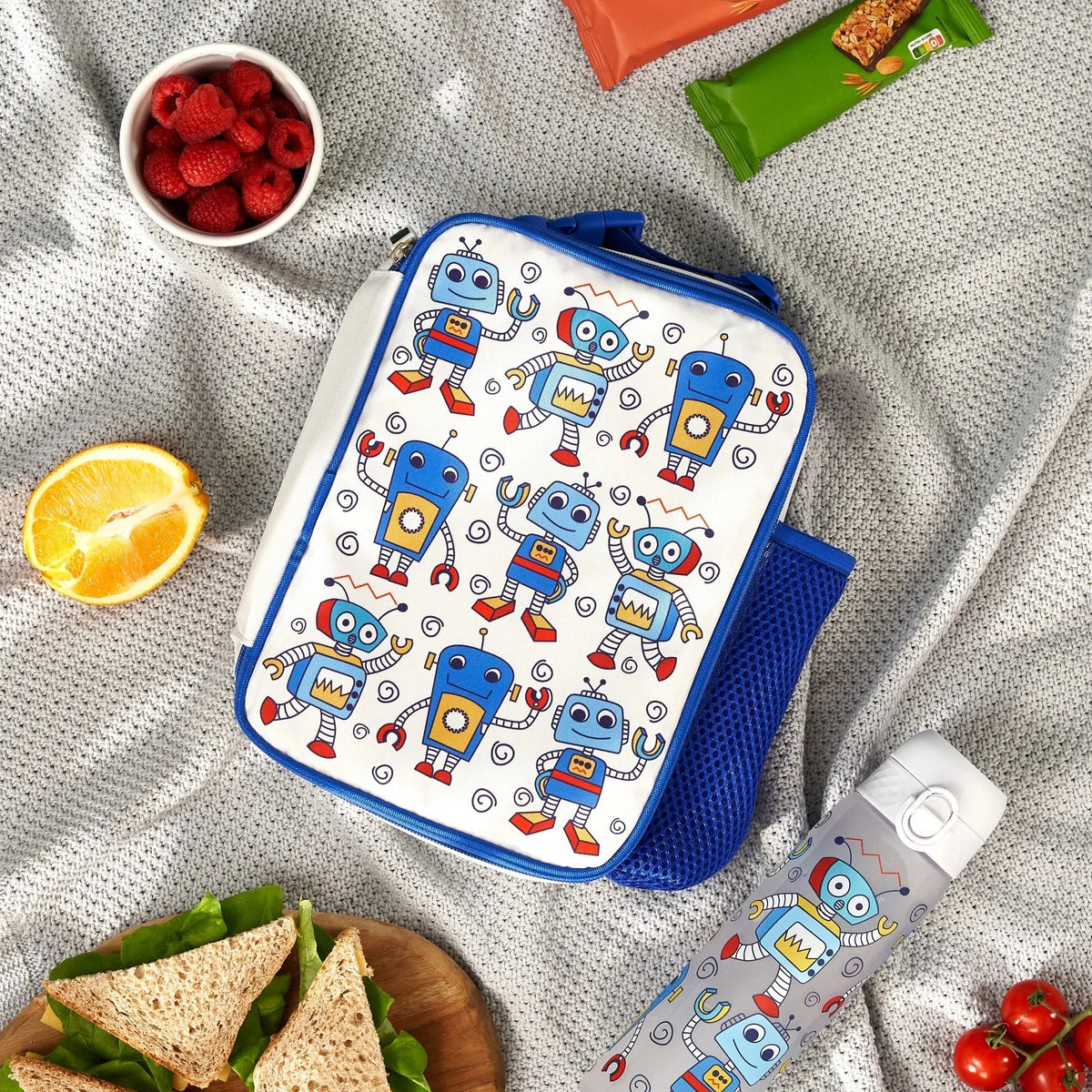 Lunchbox with cartoon robots resting on a picnic blanket surrounded by snacks including raspberries, an orange slice, sandwiches, granola bars, and a matching thermos featuring the same robot pattern.