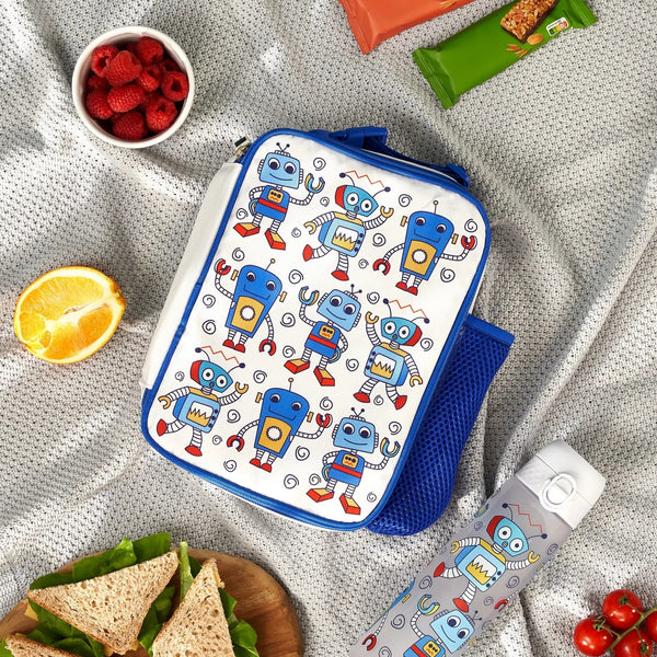 Lunchbox with cartoon robots resting on a picnic blanket surrounded by snacks including raspberries, an orange slice, sandwiches, granola bars, and a matching thermos featuring the same robot pattern.