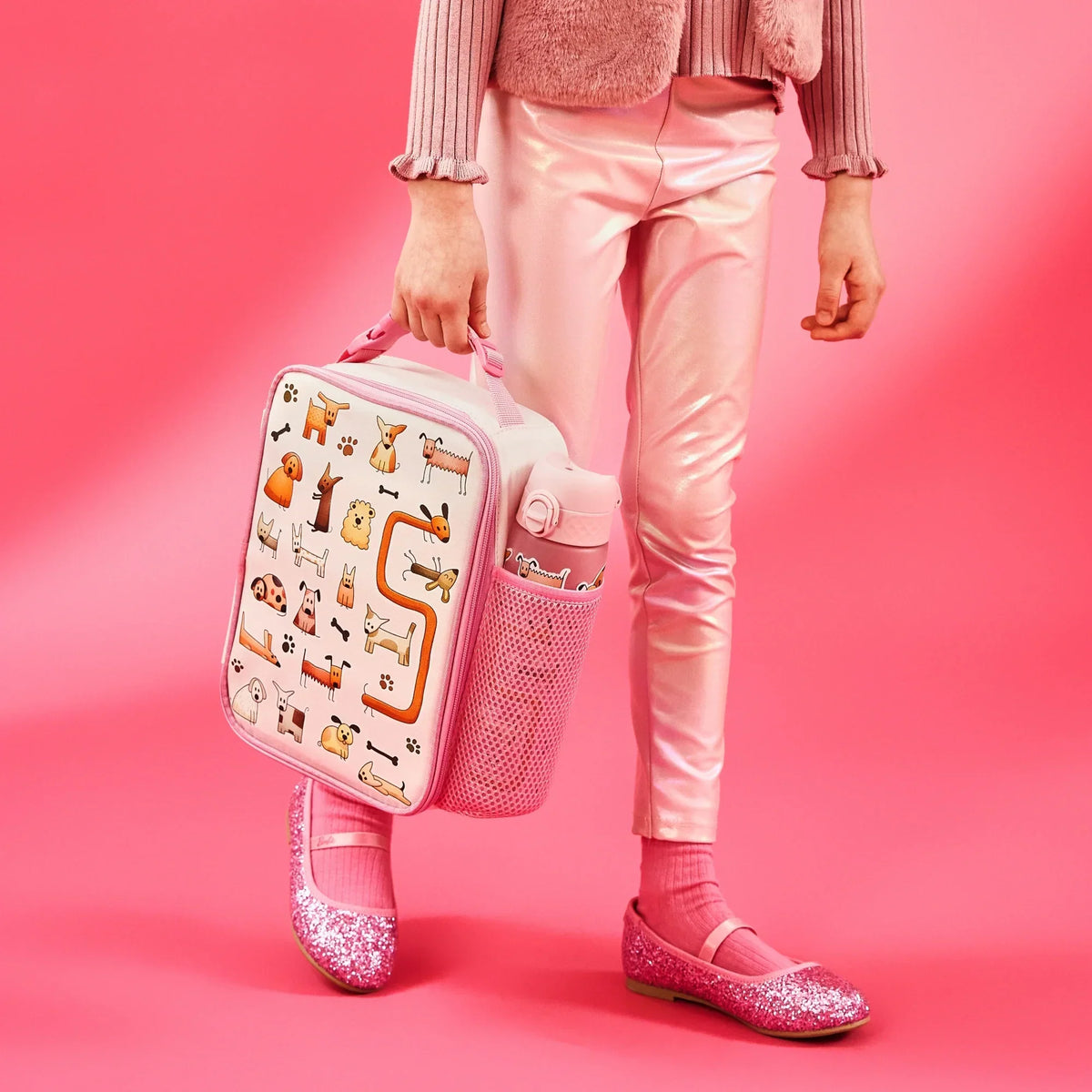 Person carrying a pink lunchbox decorated with cartoon dogs walking in a pink outfit including shiny leggings and glittery shoes against a pink background