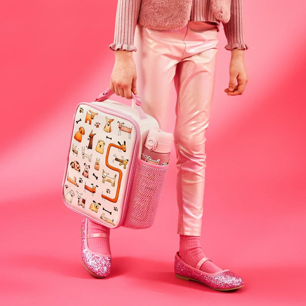 Person carrying a pink lunchbox decorated with cartoon dogs walking in a pink outfit including shiny leggings and glittery shoes against a pink background
