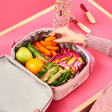 Lunchbox filled with green apple, orange, sandwich, granola bars, carrots, and Walkers crisps. A hand reaches for carrots. Nearby is a decorated water bottle on a pink table. The name 