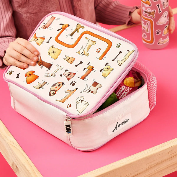 Lunchbox featuring cartoon animal designs is being opened by child wearing pink sweater at table; contains snacks with visible text: "Prawn Cocktail" and "Ion8" labeled with "Amelia."