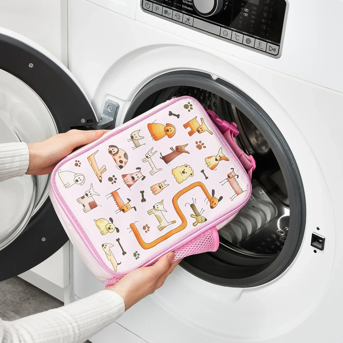 A person places a pink pet-themed zippered pouch with animal illustrations into a front-loading washing machine in a laundry room setting.