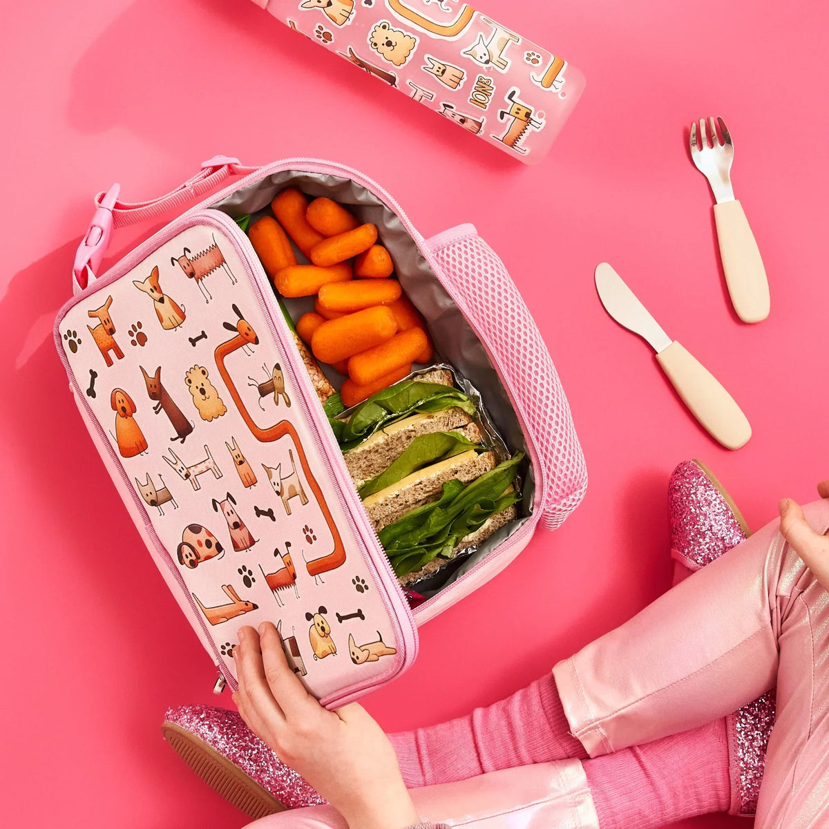 Lunchbox with cartoon dogs holds carrots and sandwiches inside vibrant pink setting with utensils and child wearing pink sparkly shoes.
