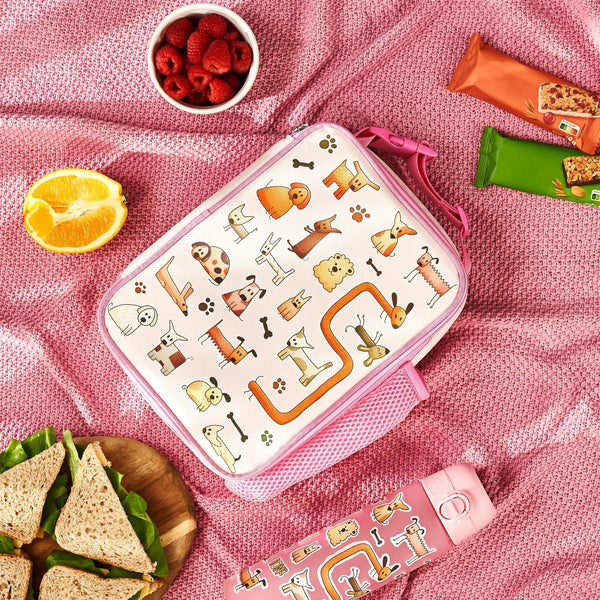 Lunchbox decorated with cartoon dogs is resting on a pink blanket, accompanied by a bowl of raspberries, orange half, sandwich plate, two snack bars, and a dog-themed bottle.