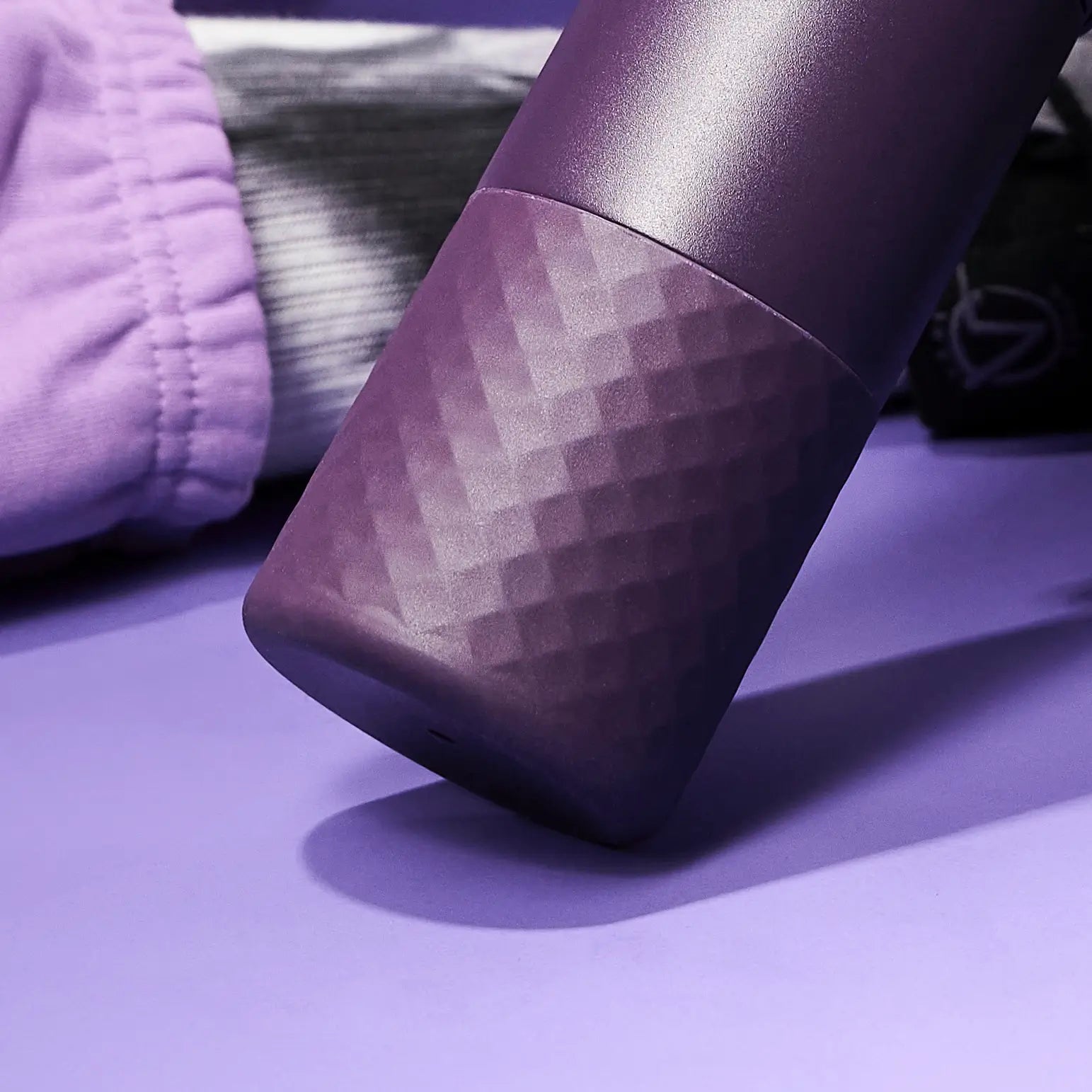 A dark purple, textured tumbler rests on a light purple surface near purple and gray fabric.
