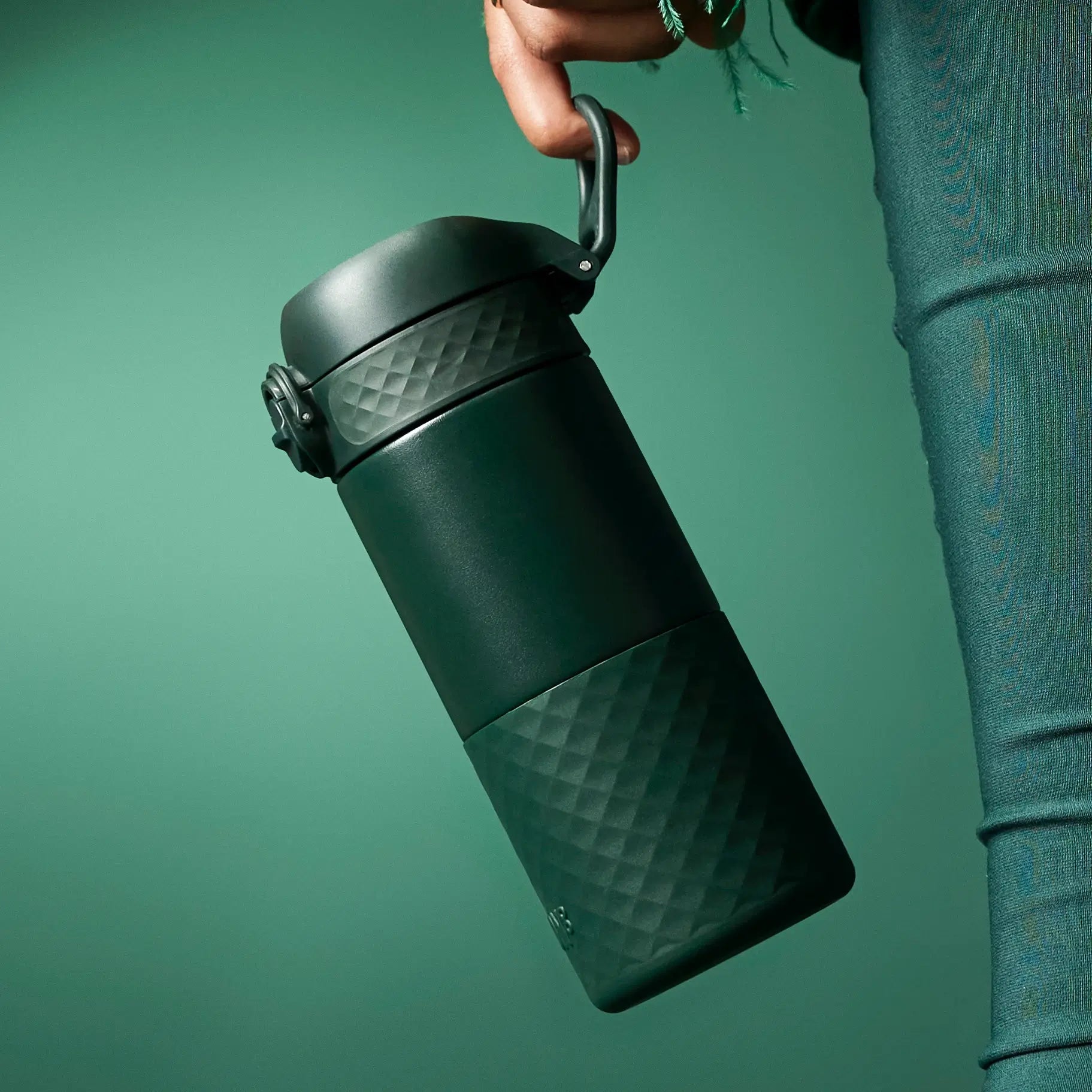 A dark-green thermos is being held by a hand. The thermos has a textured, diamond pattern and a carrying loop. The background is a matching green.