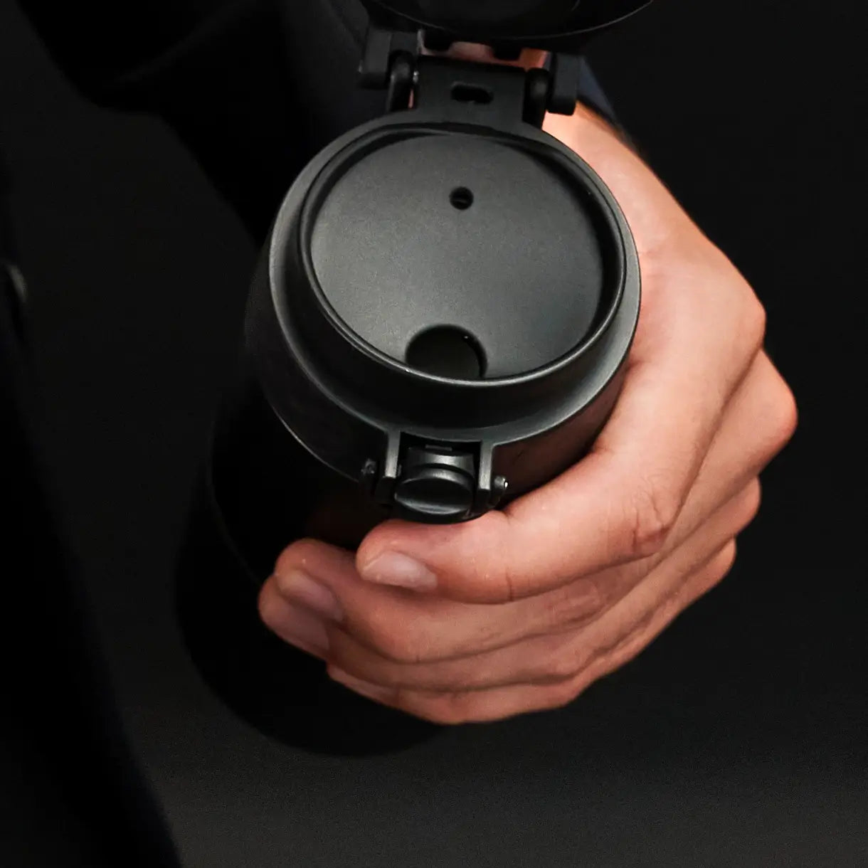 A black travel mug is held; its lid is closed. The hand grips the mug in a dark setting.