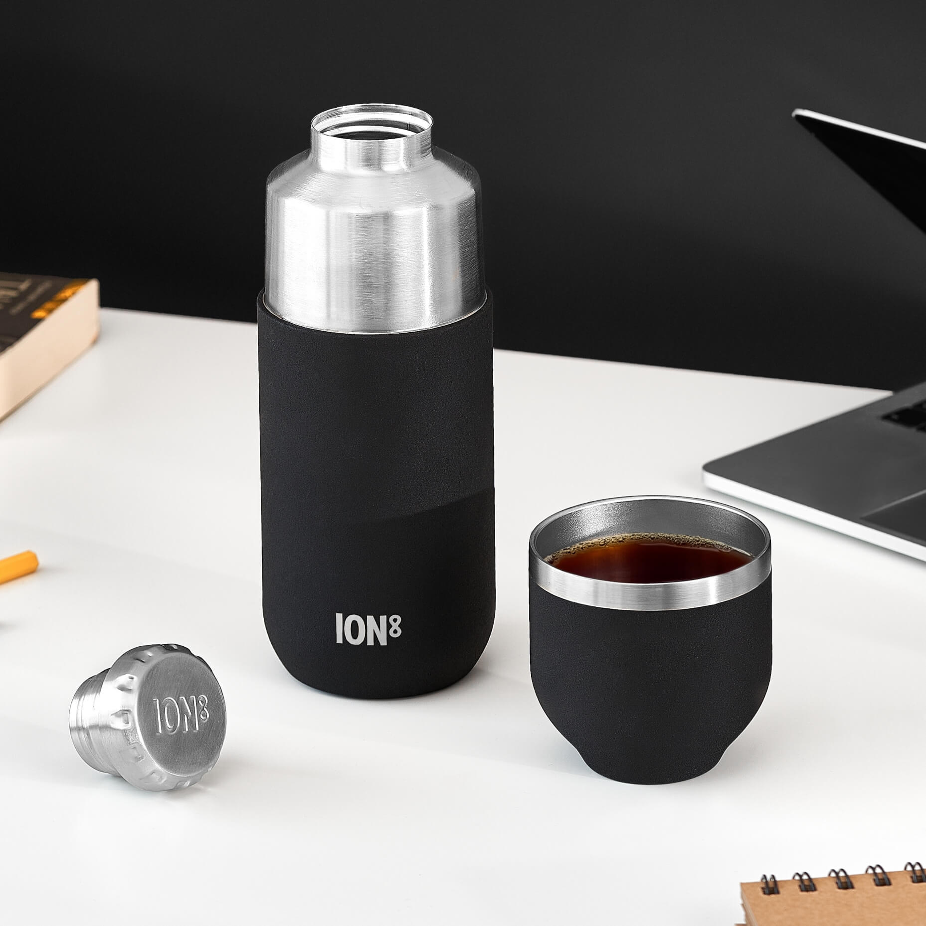 A black and silver ION8 thermos sits on a white desk, next to a cup of dark liquid and a laptop. The thermos has a black sleeve and the text "ION8" is visible on it. A silver ION8 cap is also present.