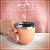 A hand holds an insulated 300ml/10oz cup with a metallic rim on a table against a pink garment background. Text: 