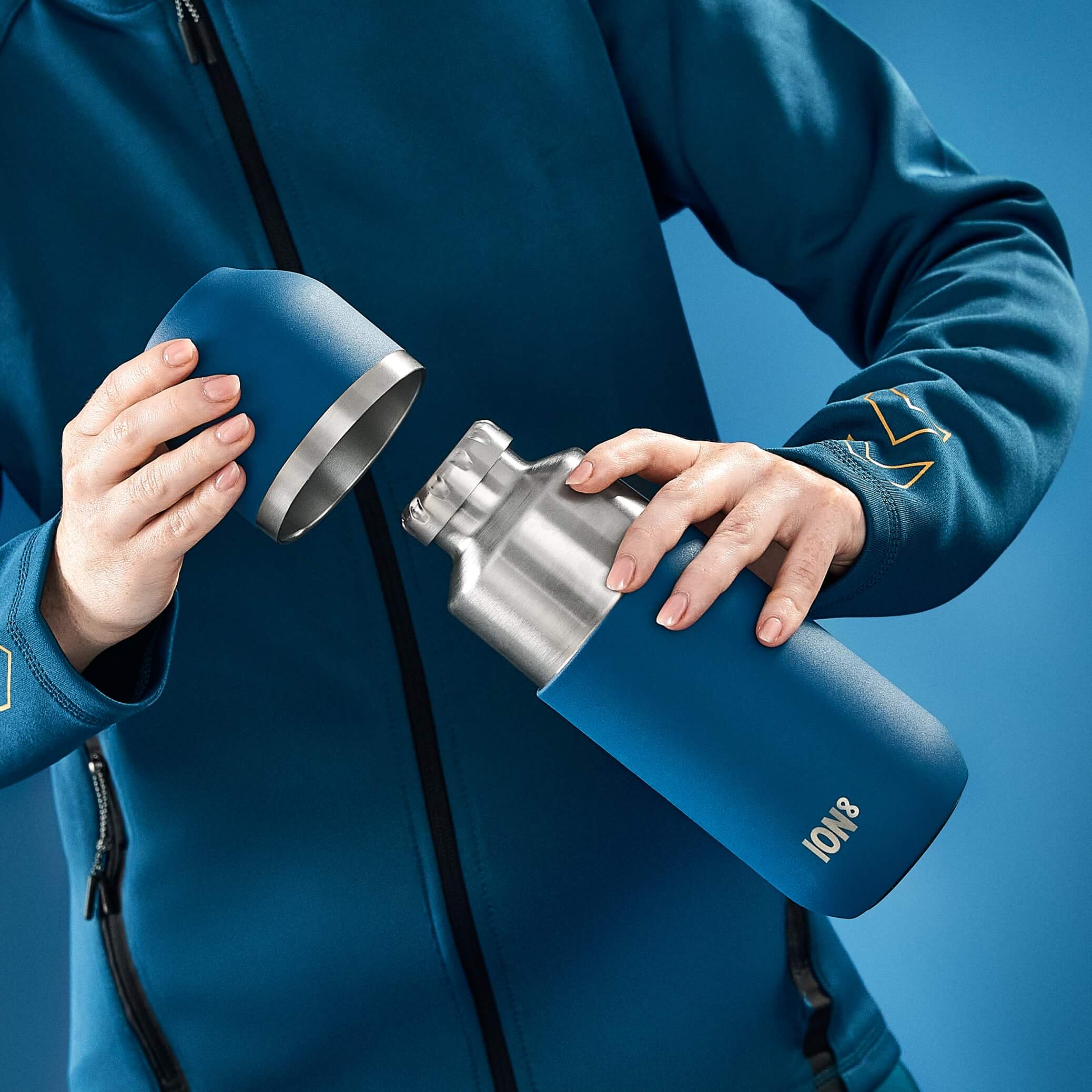 A person opens a blue ION8 insulated water bottle against a blue background. The bottle is stainless steel with a blue sleeve.