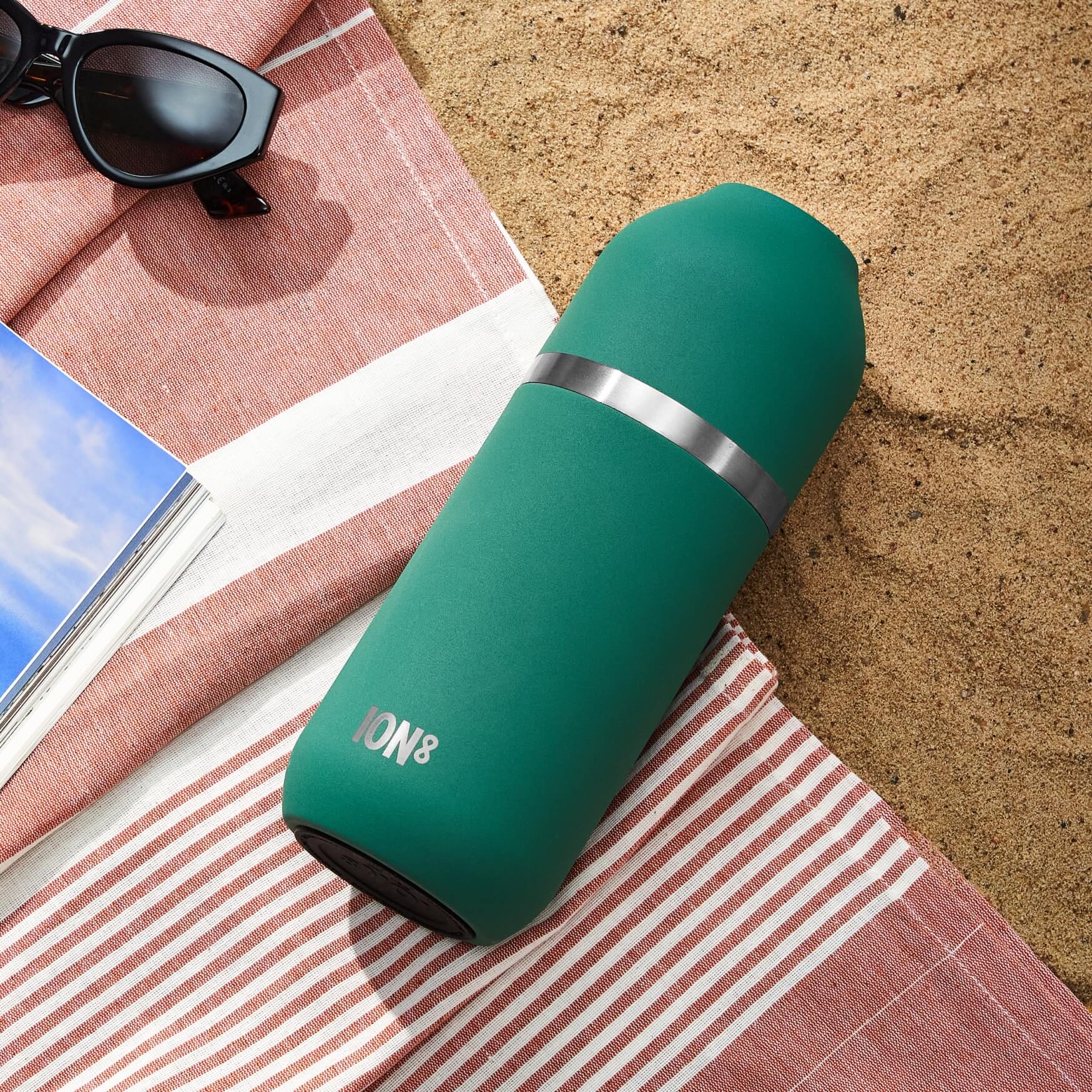 A teal ION8 thermos rests on a red and white striped beach towel on sand.