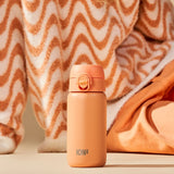 Orange water bottle standing on a beige surface ION8 printed at the bottom surrounded by wavy patterned and solid fabrics in warm tones