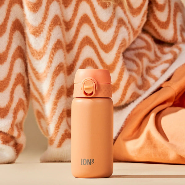 Orange water bottle standing on a beige surface ION8 printed at the bottom surrounded by wavy patterned and solid fabrics in warm tones