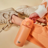 A hand holds an orange Ion8 water bottle near crossed legs wearing peach pants and beige sneakers on a light beige surface