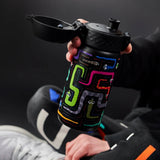 Hand holds a colorful, patterned water bottle with an open lid showing a zigzag design and cartoon eyes in a dark setting. A person wears black clothing and white sneakers nearby.