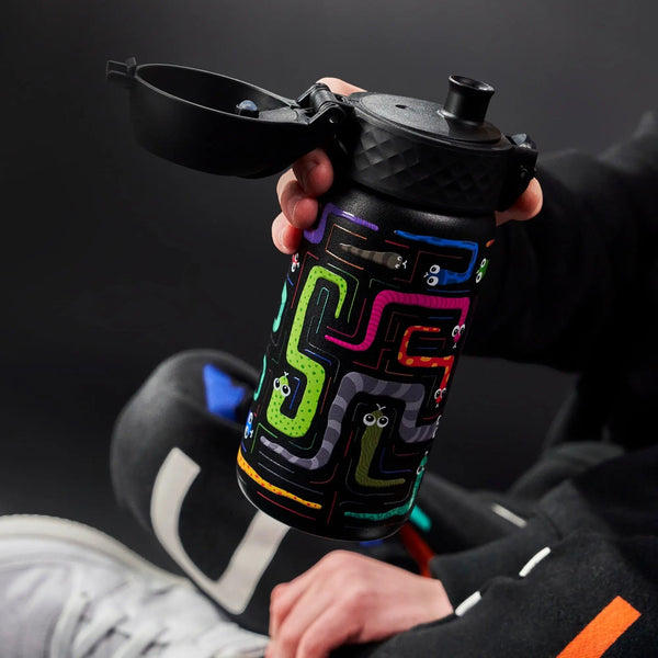 Hand holds a colorful, patterned water bottle with an open lid showing a zigzag design and cartoon eyes in a dark setting. A person wears black clothing and white sneakers nearby.