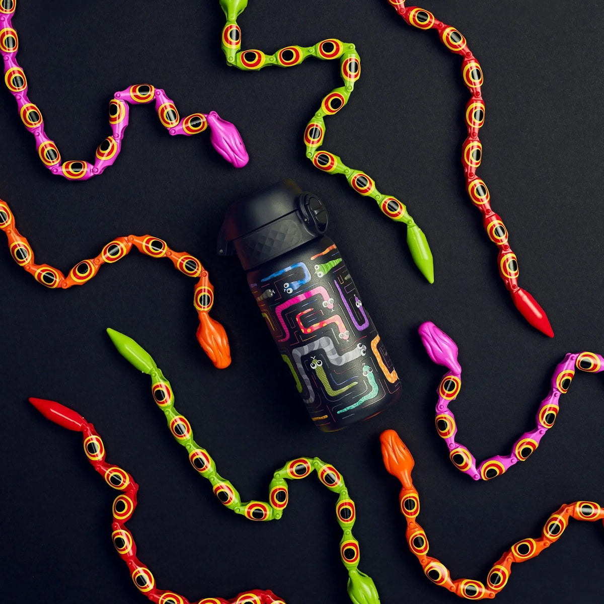 A black water bottle, decorated with colorful, playful snake designs, sits on a dark background surrounded by multicolored toy snakes in vibrant red, orange, pink, and green.