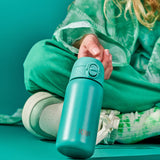 A turquoise water bottle is held by a person in green tie-dye clothing sitting cross-legged against a matching teal background. Text on the bottle reads ION8.