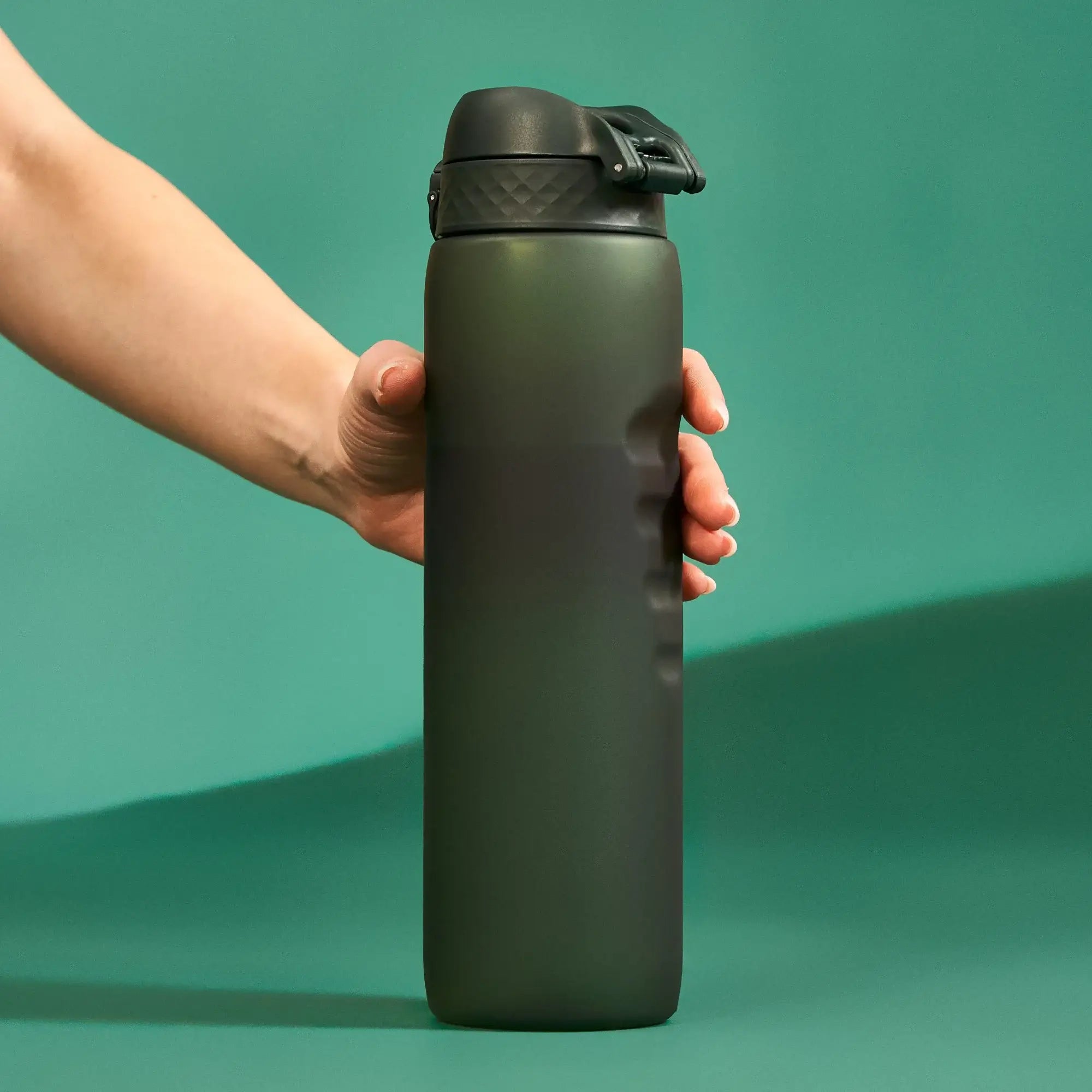 A dark-green water bottle is held by a hand against a teal background.