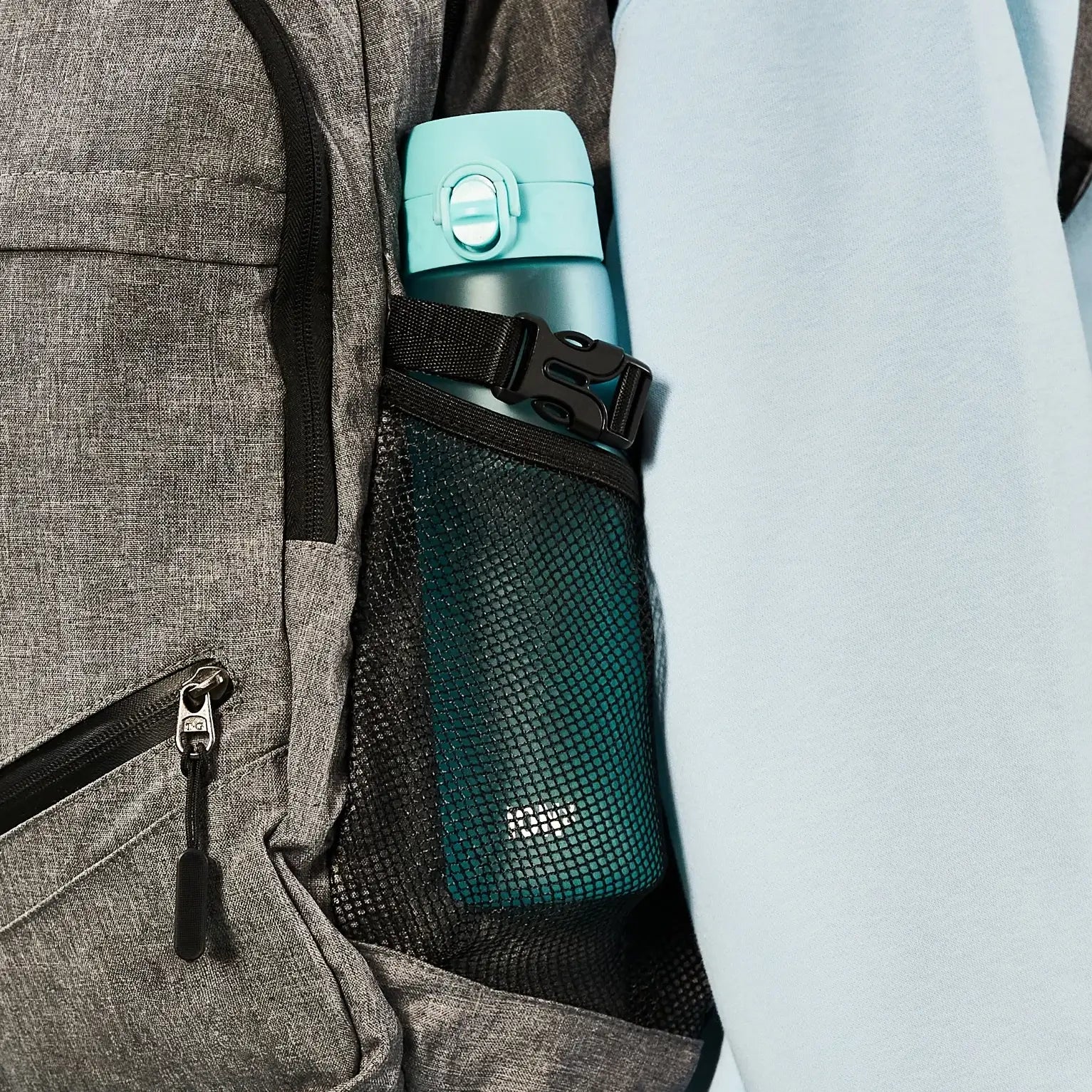 A teal water bottle is secured in a mesh pocket of a gray backpack. The backpack's side is adjacent to a light-blue garment.