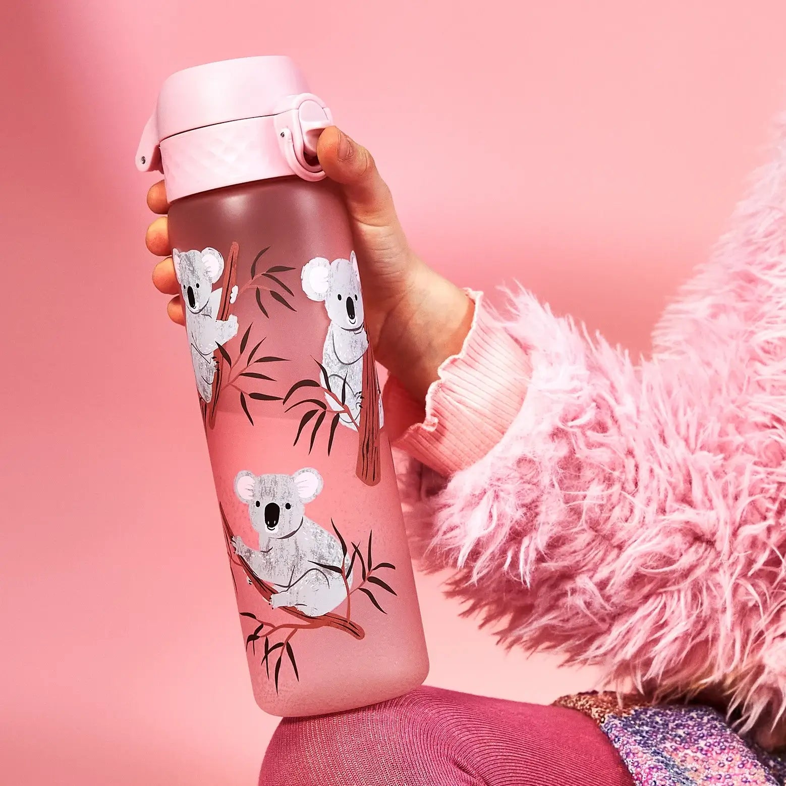 A pink water bottle, featuring illustrated koalas on branches, is held by a person wearing a fluffy pink sweater against a pink background.