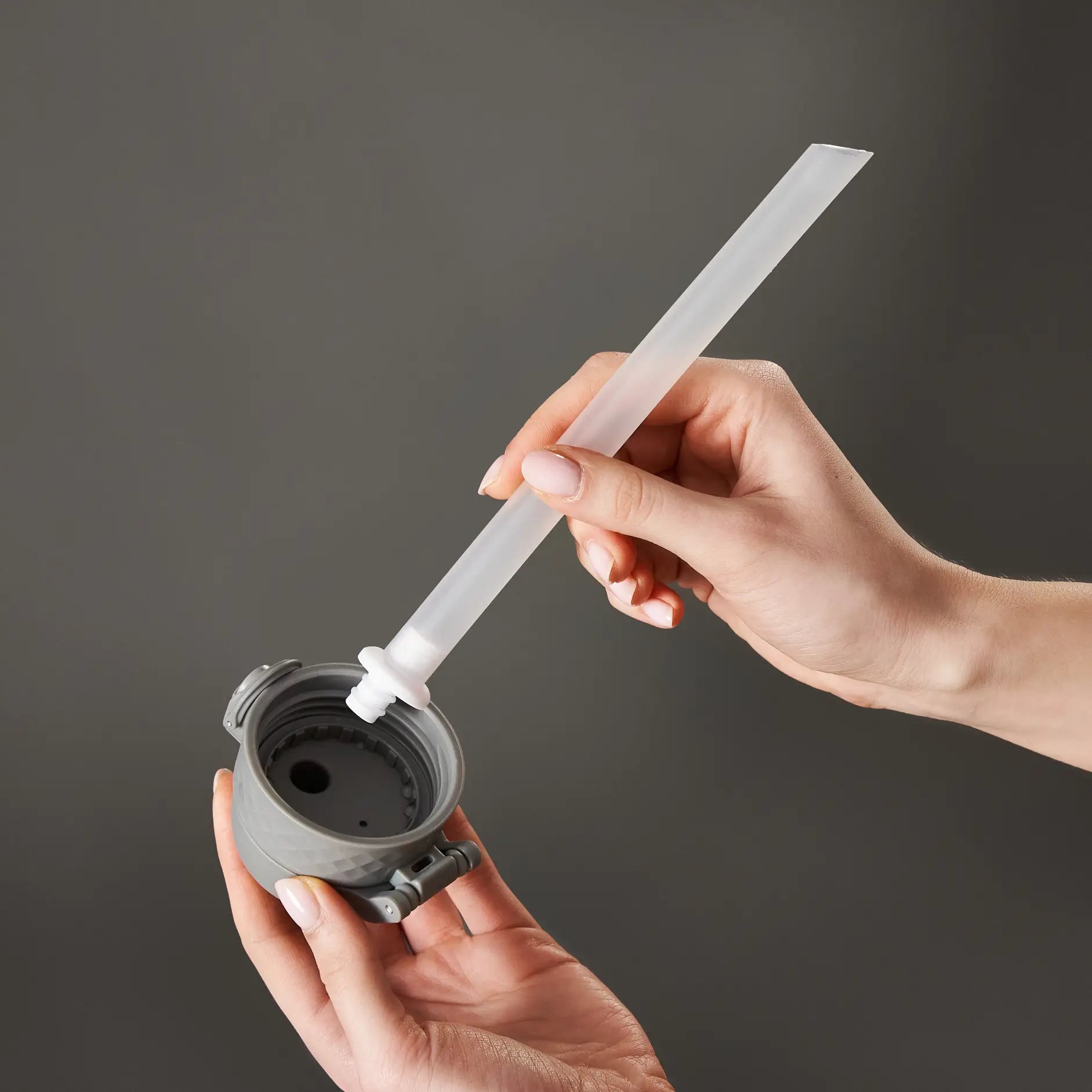 A straw is being inserted into a gray bottle cap. The action is taking place against a dark gray background.