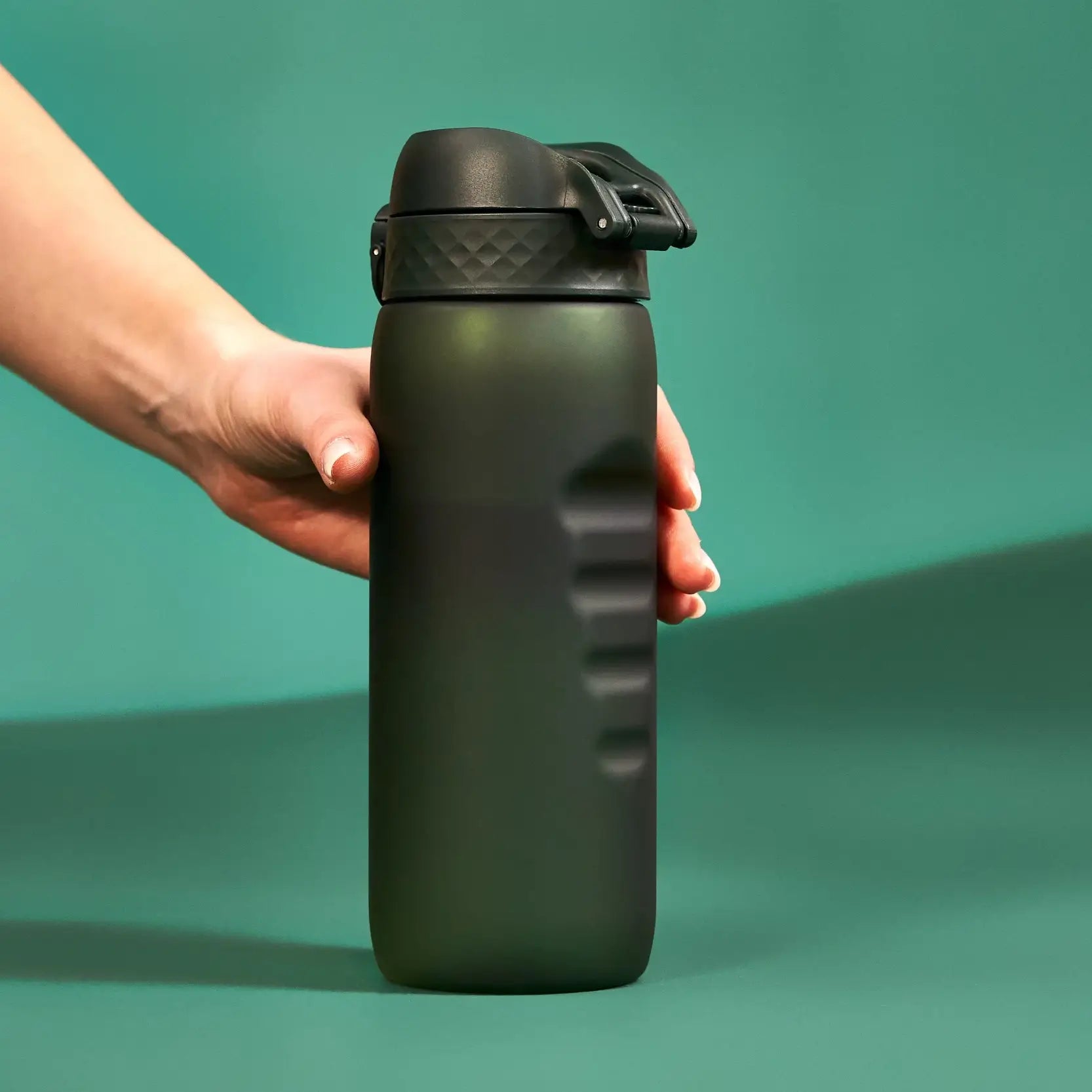A dark-green water bottle with a black lid is held by a hand against a teal background.