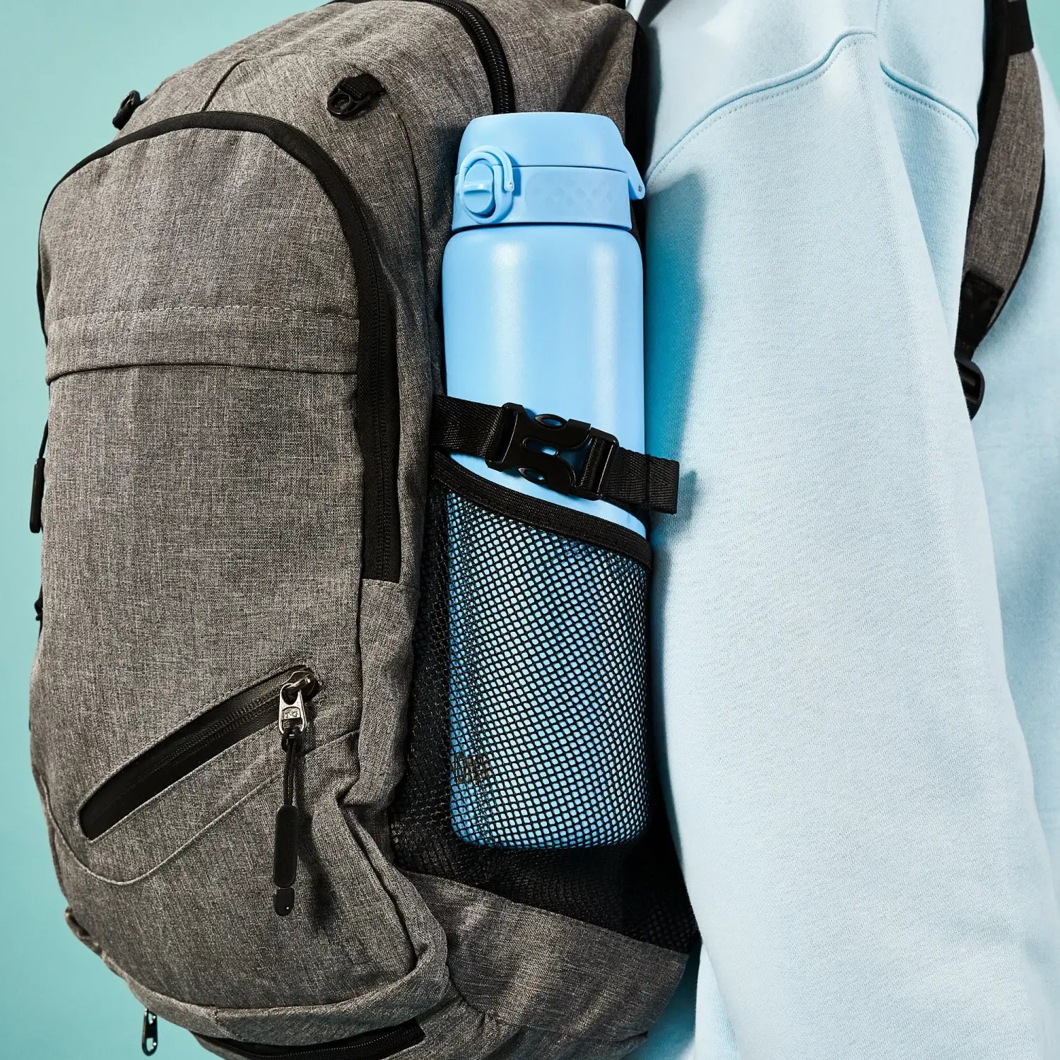 A light blue water bottle is secured in a grey backpack's side pocket, worn by a person in a light blue sweatshirt against a teal background.