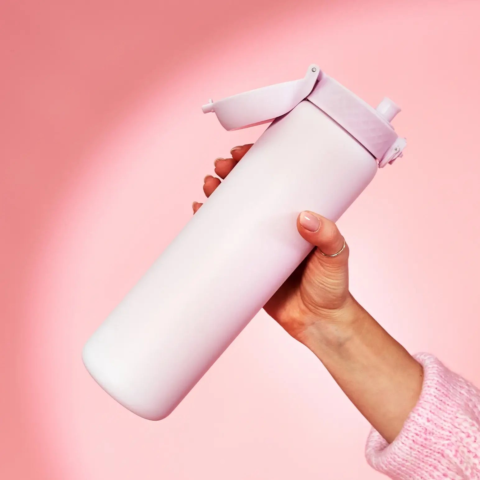 A pale pink water bottle is held by a hand; the bottle’s flip top is open. The background is a solid pink.