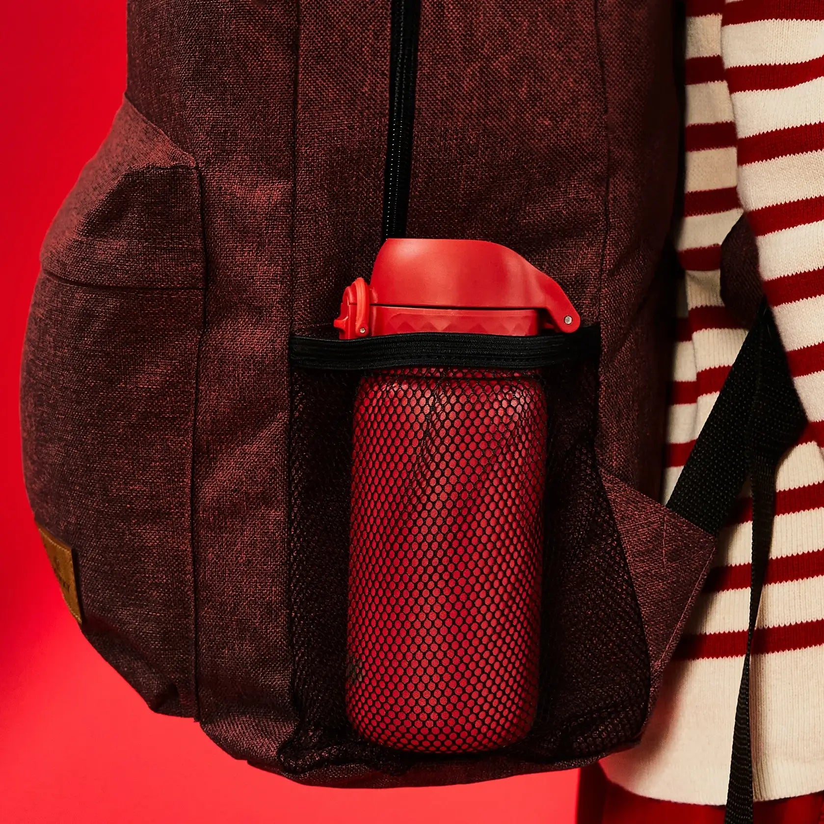 A red water bottle rests in a maroon backpack's mesh pocket. The backpack is being worn against a solid red background.