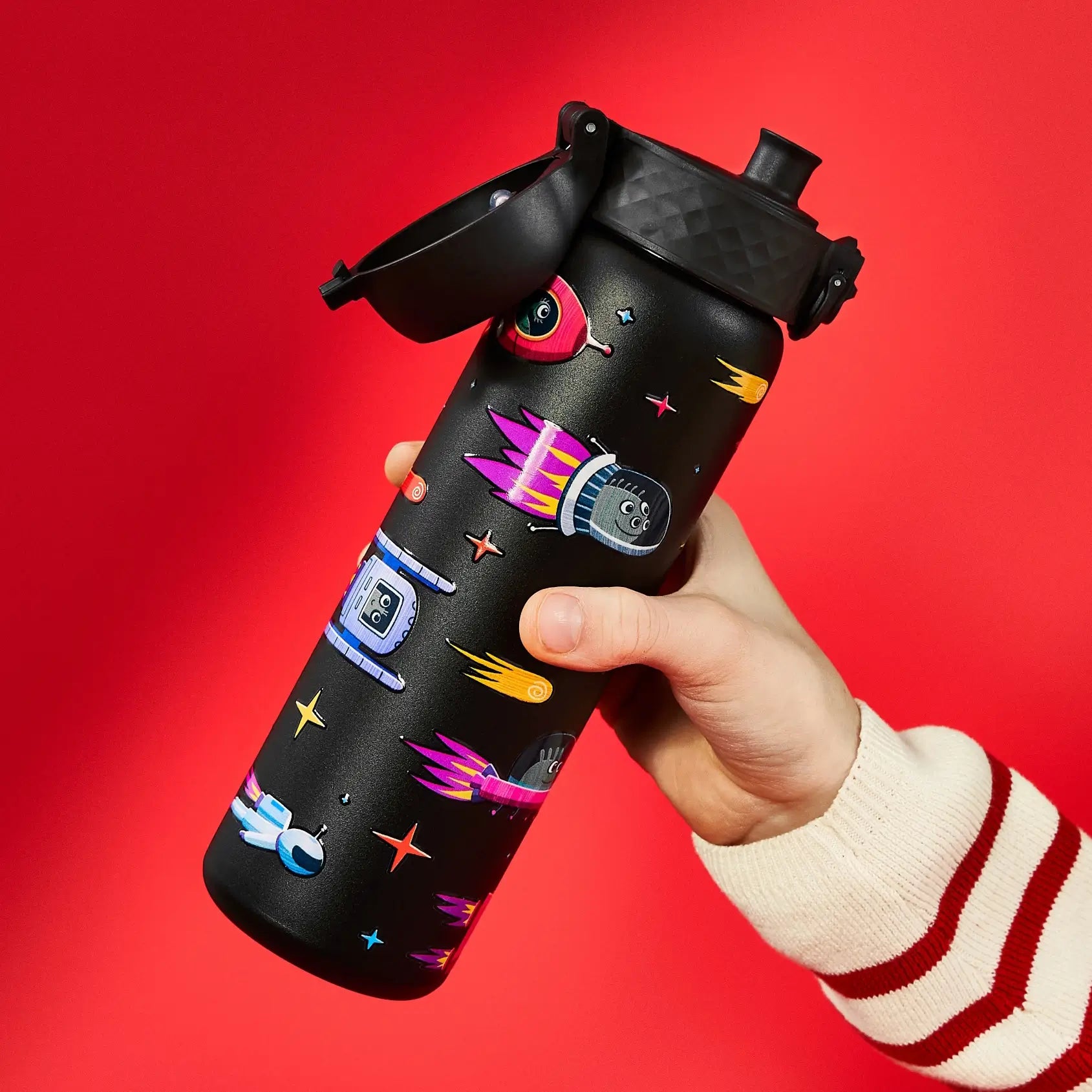 A black water bottle, decorated with space-themed illustrations, is held in a hand against a red background. The bottle's lid is ajar.