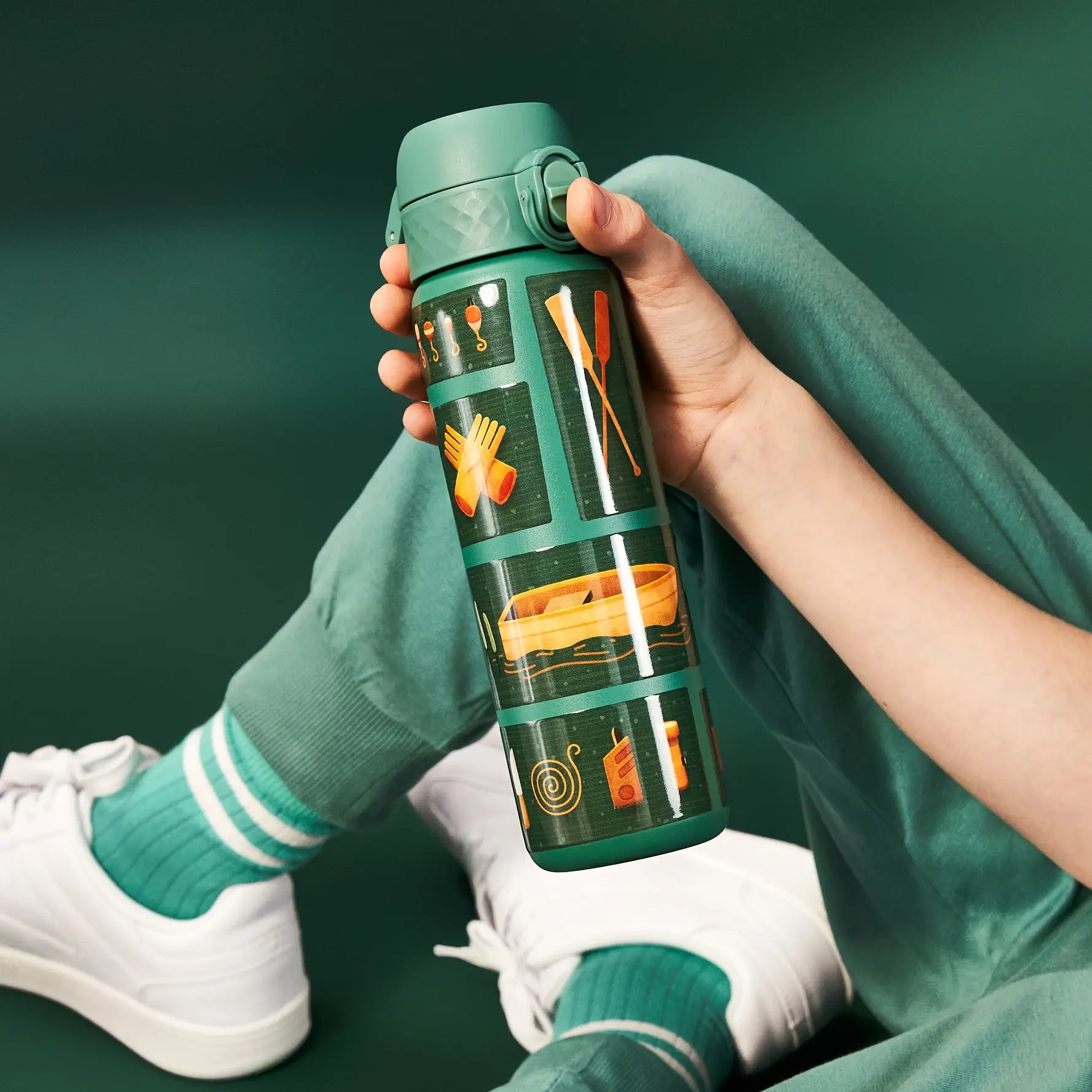 A person holds a green water bottle with illustrated outdoor scenes. The bottle is decorated with images of a boat, oars, gloves, and other camping items. The setting is a dark green background.