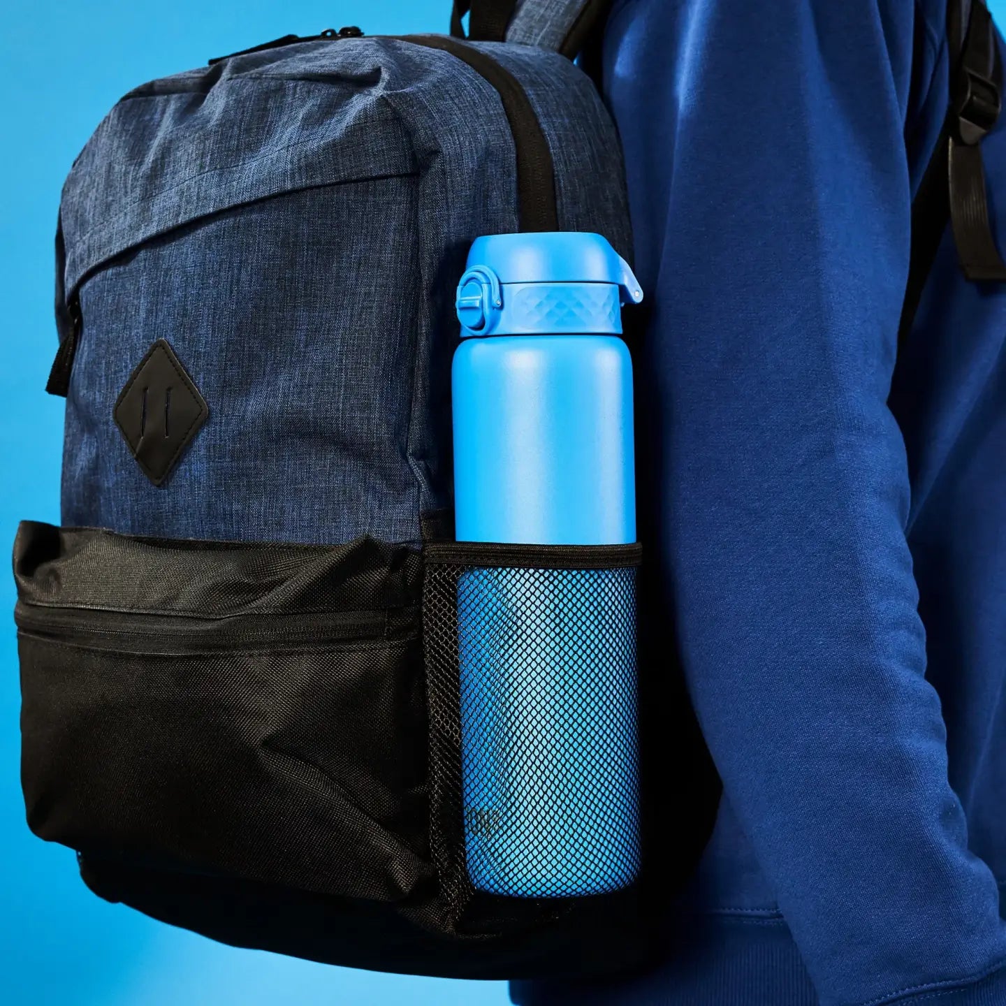 A blue backpack holds a blue water bottle in a mesh side pocket. The backpack is worn by a person in a blue sweatshirt against a blue background.